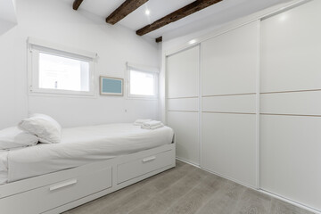 Youth bedroom with bed with white linens, two small windows in the wall, drawers in the bed and a large built-in wall-to-wall wardrobe with sliding doors