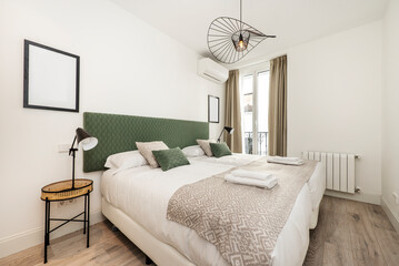 Bedroom with large bed with white sheets, green cushions, aluminum radiator, round bedside tables, balcony with curtains and brown parquet floor