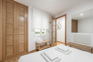 Bedroom with large bed with white sheets, clean white towels, frameless mirror on the wall, built-in wardrobe with wooden doors and matching benches