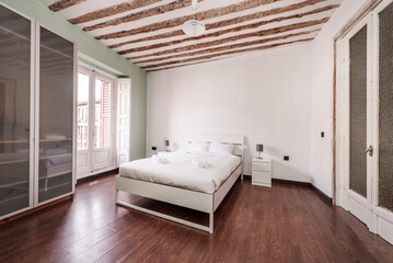 Naklejka na ściany i meble Bedroom with large bed with white sheets, clean white towels, wardrobe with glass doors, exposed wooden beams on the ceiling and balcony with white wooden shutters