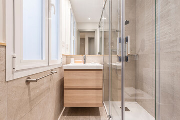 Bathroom with white aluminum window, frameless mirror embedded in the wall, hanging wooden cabinet and white porcelain sink and shower cabin with glass partition