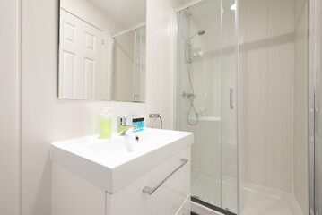 Fototapeta na wymiar Bathroom with white walls and frameless wall mirror, white porcelain sink on dresser and walk-in shower