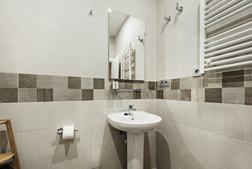 Bathroom with cream-colored tiles and dark-colored border, frameless wall mirror, white porcelain sink on matching pedestal and white heated towel rail