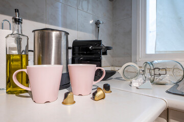 Corner of a kitchen with a pod coffee machine next to pale pink mugs next to a stainless steel kettle and oil can