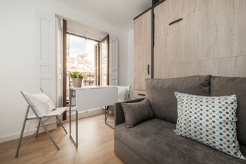 Extra bed inside sofa with cushions in short-term rental studio apartment with wardrobe, white wooden folding table with chairs and light wooden floor and wooden shuttered balcony