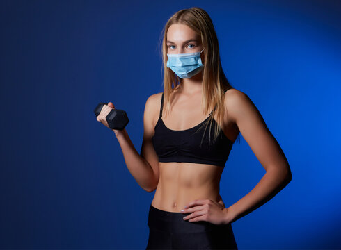 A fit woman in a face mask to avoid the spread of coronavirus is doing bicep curls with dumbbells.