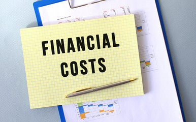 FINANCIAL COSTS text written on notepad with pencil. Notepad on a folder with diagrams.