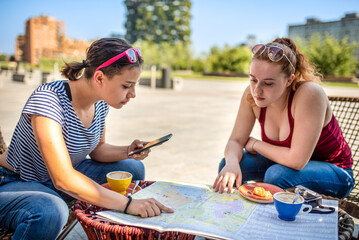young female tourists examining the map of the city they are visiting during their travel, breakfast time at the bar looking for hostel or interesting places