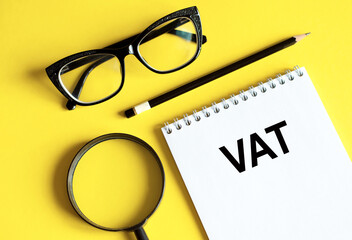 acronym VAT value added tax in notebook with pencil, magnifier, and glasses on a yellow background