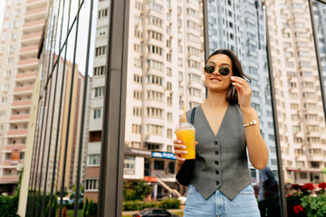 Happy lady is drinking lemonade and touching her glasses on the street. Brunette woman in light jacket and jeans with dark bag smiling and holding summer drink outside.