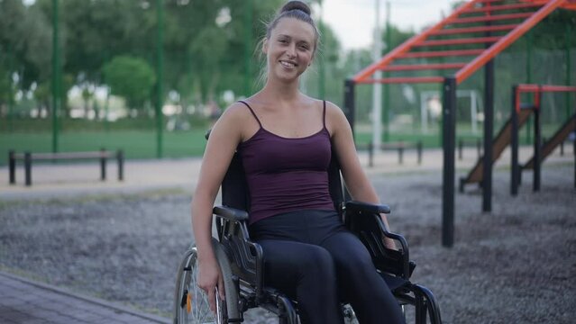 Young positive woman in wheelchair smiling looking at camera sitting on sports ground in park. Portrait of happy confident Caucasian sportswoman with disability posing in slow motion outdoors