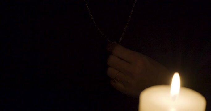 Hand clasping silver crucifix hanging around neck in a dark room with a burning candle in the foreground