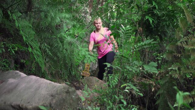 Woman in pink shirt walking on trail, going over a rock in tropical forest.
