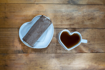 A white cup of black strong coffee and cream ice cream in chocolate in a heart-shaped mug and plate in the shape of a heart. Hot drink and dessert on brown wooden background.