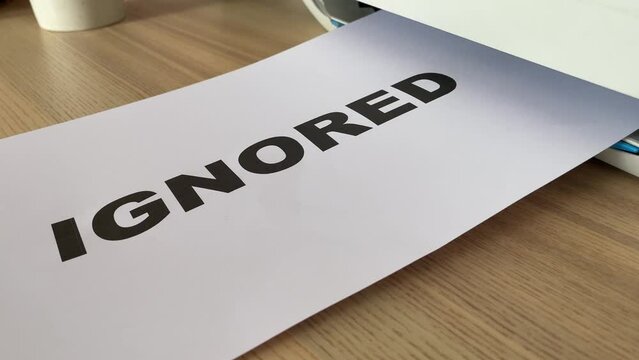 Printing &ldquo;IGNORED&rdquo; black text written message on paper sheet document. Close up A4 page is being printed from multifunction printer scanner on wooden desk in office. Male office worker hand take paper