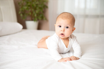 Portrait of a cute baby girl of 5 months in a white bodysuit on a white bed in the bedroom. Space for text. Child lifestyle