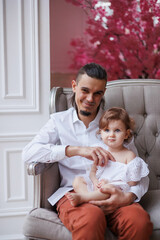 Fototapeta premium Handsome happy father sitting with his little daughter on beige sofa indoors. Young man in white shirt with his 1-year-old daughter in white dress, holding her in hands on legs. family tenderness