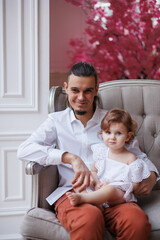 Obraz premium Handsome happy father sitting with his little daughter on beige sofa indoors. Young man in white shirt with his 1-year-old daughter in white dress, holding her in hands on legs. family tenderness