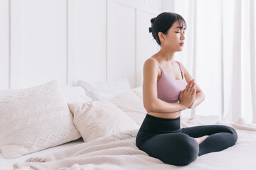 Young serene attractive sporty Asian woman practicing yoga on the bed, doing Ardha Padmasana exercise, meditating in Half Lotus pose with namaste, indoor working out at home, wearing sportswear.