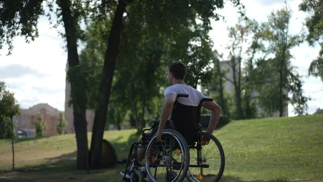 Tracking shot side view of young man pushing wheelchair wheels riding fast in sunbeam. Determined motivated Caucasian sportsman training exercising outdoors in park. Disability and sport concept