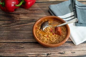 top view of an earthenware dish with lentils with rice and vegetables with chorizo and blood sausage on a wooden table with red peppers and a tea towel