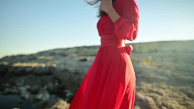 Close-up of a woman in a flattering red dress walking on a sea rocky landscape. Slow motion model with golden sunset illumination. slim model close-up dress in the wind