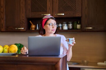 Middle aged woman in home kitchen relaxing using laptop for leisure and entertainment