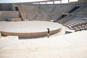 Man in a smart casual, with a hat and looking far, is standing outdoors in an amphitheater