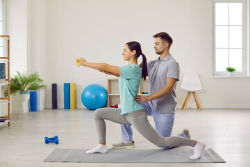 Male physiotherapist helping his female patient do special physical exercises. Side view of happy young woman doing forward lunges on rubber mat at physiotherapy clinic or rehabilitation center - Powered by Adobe