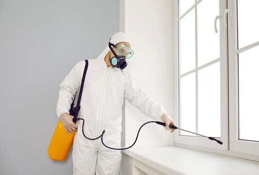 Pest control exterminator working inside house. Young guy wearing protective white suit and mask holding sprayer bottle and spraying insecticide over window sill in modern living room interior at home