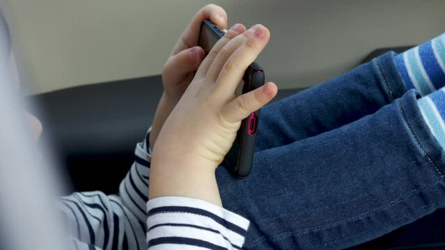 cute little boy is smiling and sitting on a modern car seat, with phone in his hands. handsome brunnete kid in stripped shirt and blue jeans. phone addicted child. young blogger, ignoring parents. 