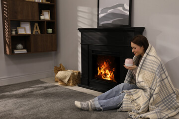 Beautiful young woman with cup of hot drink sitting on floor near fireplace at home. Space for text