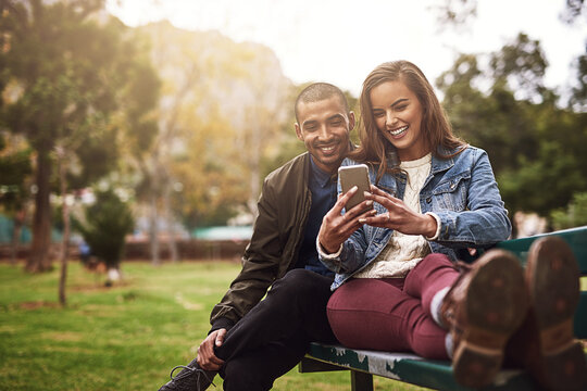 What Do You Think About This Photo. Shot Of A Cheerful Young Couple Sitting Down On A Bench While Using A Phone Together Outside In A Park.