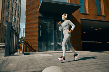 Full length shot of female athlete in sportswear, headphones and medical mask jogging in the city