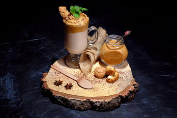 Rustic image of glass of dalgona coffee and ingredients on woody stand in dark environment