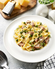 Farfalle with green peas, turkey ham and parmesan in cream sauce in a white plate on the kitchen table. Traditional Italian pasta on a light culinary background	