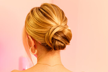 The concept of style, style and beauty. An isolated shot of a girl from behind with blonde hair gathered in a neat bun at the back of her head, against a pink studio background. A hairstyle for girls