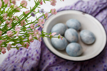 Unfocused blue painted Easter eggs in white bowl on purple kitchen table with flowers on foreground. Happy Easter