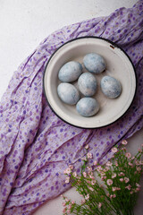 Fototapeta na wymiar Blue painted Easter eggs in white bowl on purple kitchen table with flowers. Happy Easter concept. Vertical shot, top view, copy space