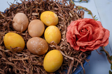 Decorative nest with painted Easter eggs and fabric rose flower on blue wooden background. Yellow and brown eggs for Easter, close up