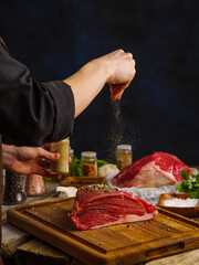 Fototapeta na wymiar The chef salts a large piece of raw meat on a wooden cutting board. Salt in frozen flight on a dark blue background. Cooking meat dishes. Recipes for restaurant and home cooking.