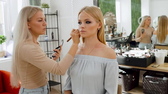 Blonde girl at a reception with a make-up artist. The make-up artist paints a brush of a lip and an eye to the woman in salon. Close-up of a make-up artist's face.
