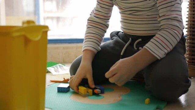 The kid assembles a construction set colorful plastic toy bricks and constructor game blocks sitting on the floor near the window. Close-up