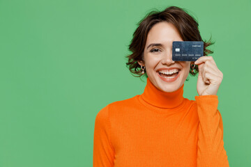 Young smiling happy cheerful fun woman 20s in orange turtleneck hold in hand cover eye with credit...