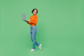 Full body young smiling woman 20s wear casual orange turtleneck hold use work on laptop pc computer...