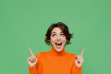 Young surprised amazed woman 20s wear orange turtleneck point index finger overhead on workspace...