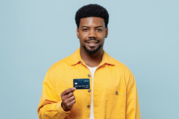 Young smiling happy fun man of African American ethnicity 20s wear yellow shirt hold in hand credit bank card isolated on plain pastel light blue background studio portrait. People lifestyle concept.