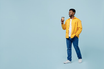 Full body young man of African American ethnicity 20s in yellow shirt hold takeaway delivery craft paper brown cup coffee to go walking isolated on plain pastel light blue background studio portrait