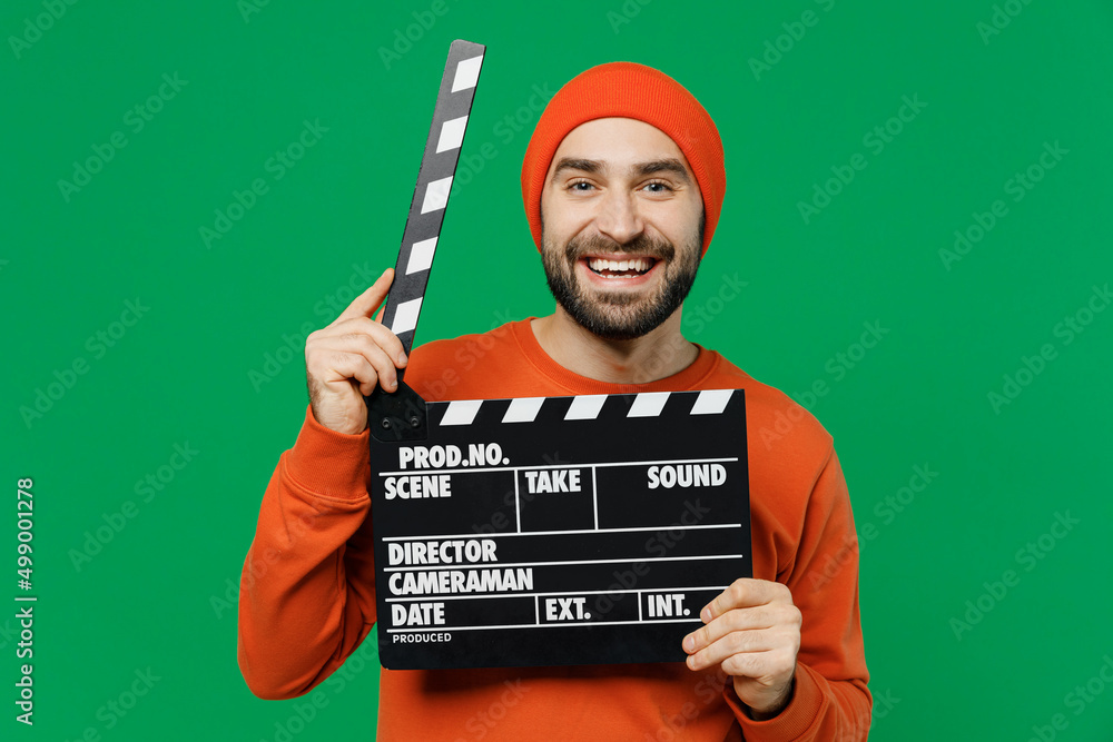 Wall mural Young smiling happy cheerful caucasian man 20s wear orange sweatshirt hat holding classic black film making clapperboard isolated on plain green background studio portrait. People lifestyle concept.