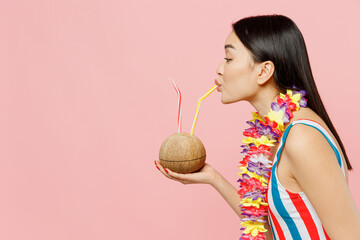 Side view young woman of Asian ethnicity in striped one-piece swimsuit hawaii lei drink coconut...